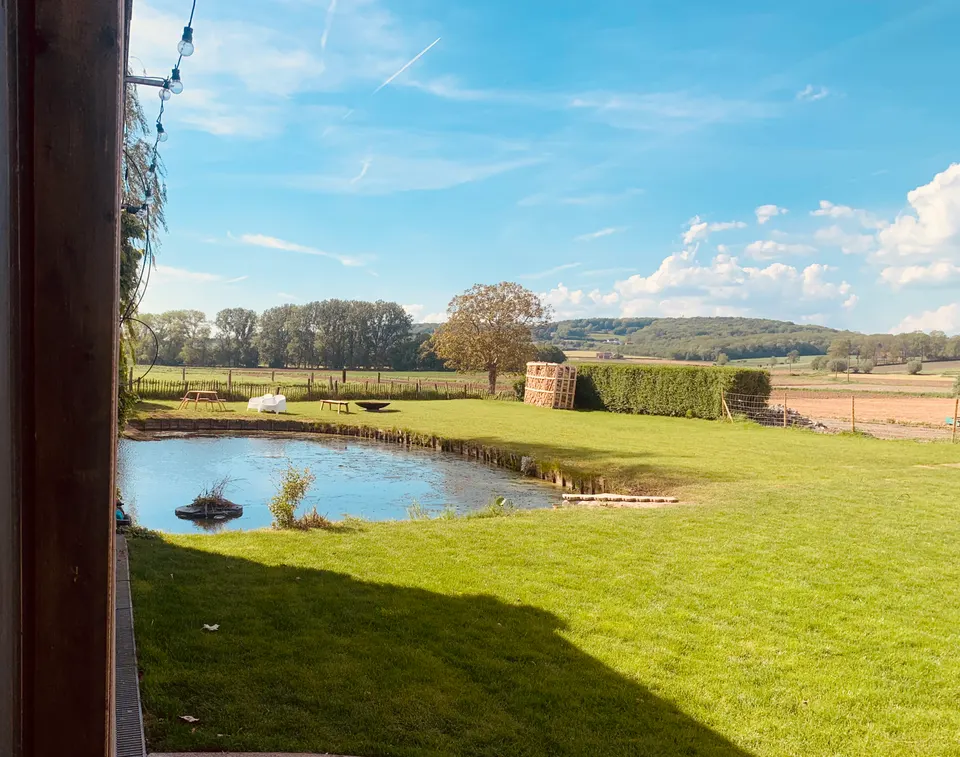 Zonnig gazon met vijver, vuurschaal en houtstapel, met akkers en beboste heuvels op de achtergrond
